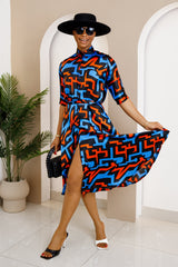 Woman wearing a vibrant Akwu shirt dress with a bold blue, black, and orange geometric print, accessorized with a black hat, sunglasses, and heels.
