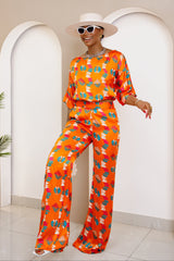 Woman wearing a vibrant orange wide-leg pant set with African-inspired prints, styled with a white hat, sunglasses, and statement accessories.