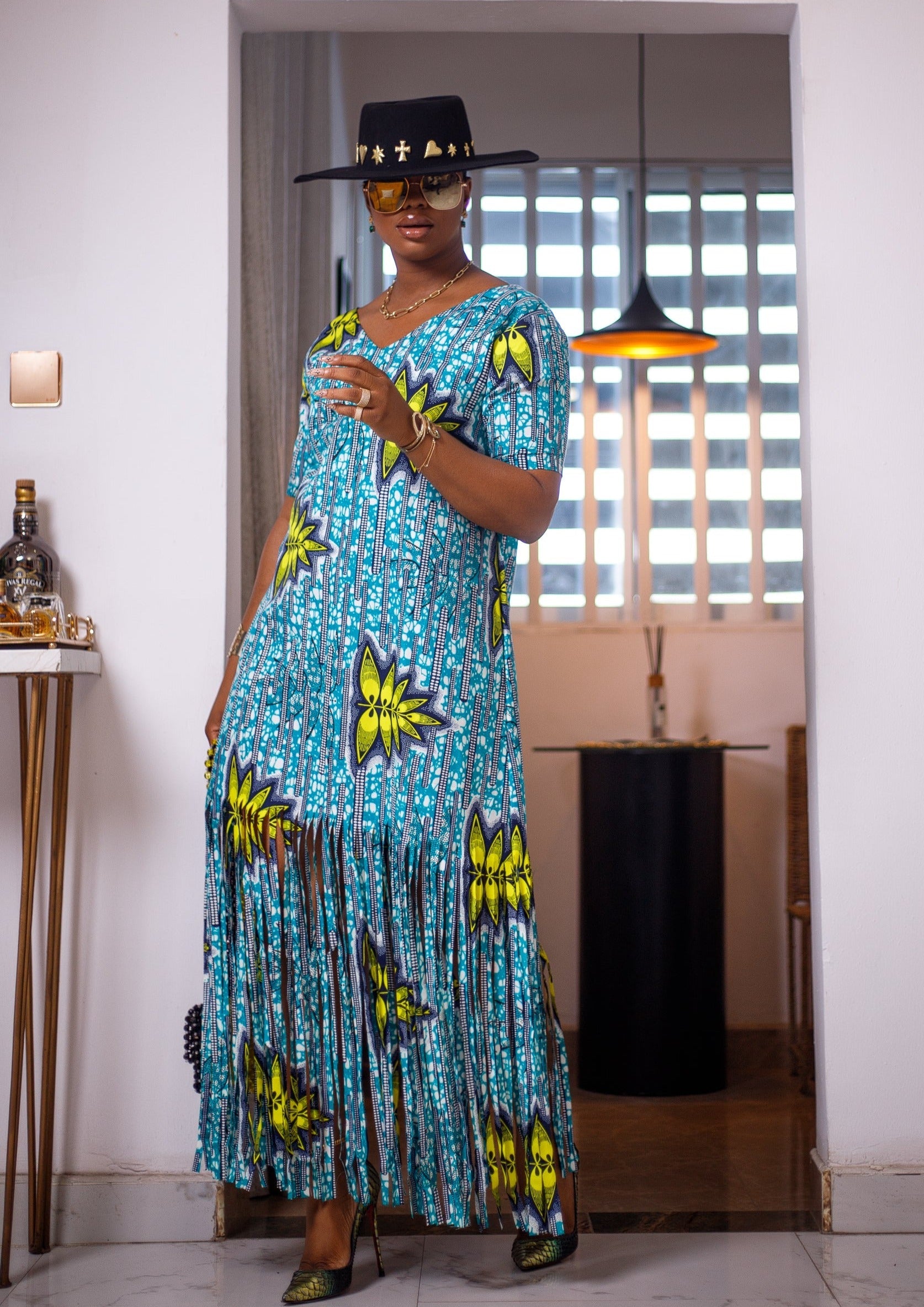 A woman posing in a blue, green, and white maxi dress with frills. She is pictured facing the camera. She is styled with a black and gold sun hat, gold jewelry, and multi-colored high heels.