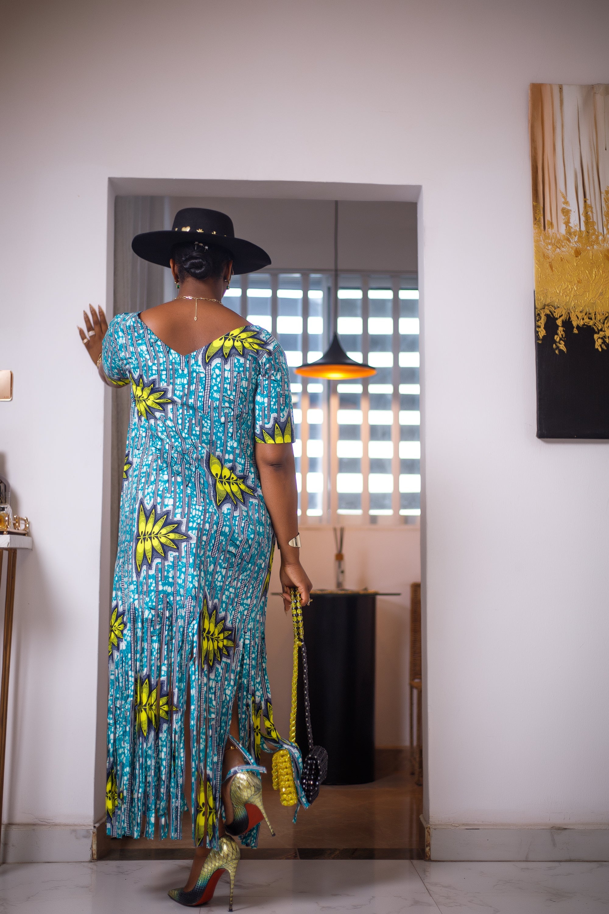 A woman posing in a blue, green, and white maxi dress with frills. She is pictured facing away from the camera. She is styled with a black and gold sun hat, a black and yellow beaded purse, and multi-colored high heels.