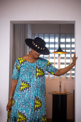A woman posing in a blue, green, and white maxi dress with frills. She is pictured from the waist up and is looking away from the camera. She is styled with a black and gold sun hat and gold jewelry.