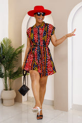 Woman wearing a vibrant mini shirt dress in red, black, and yellow geometric print, accessorized with a red hat, sunglasses, and black heels.