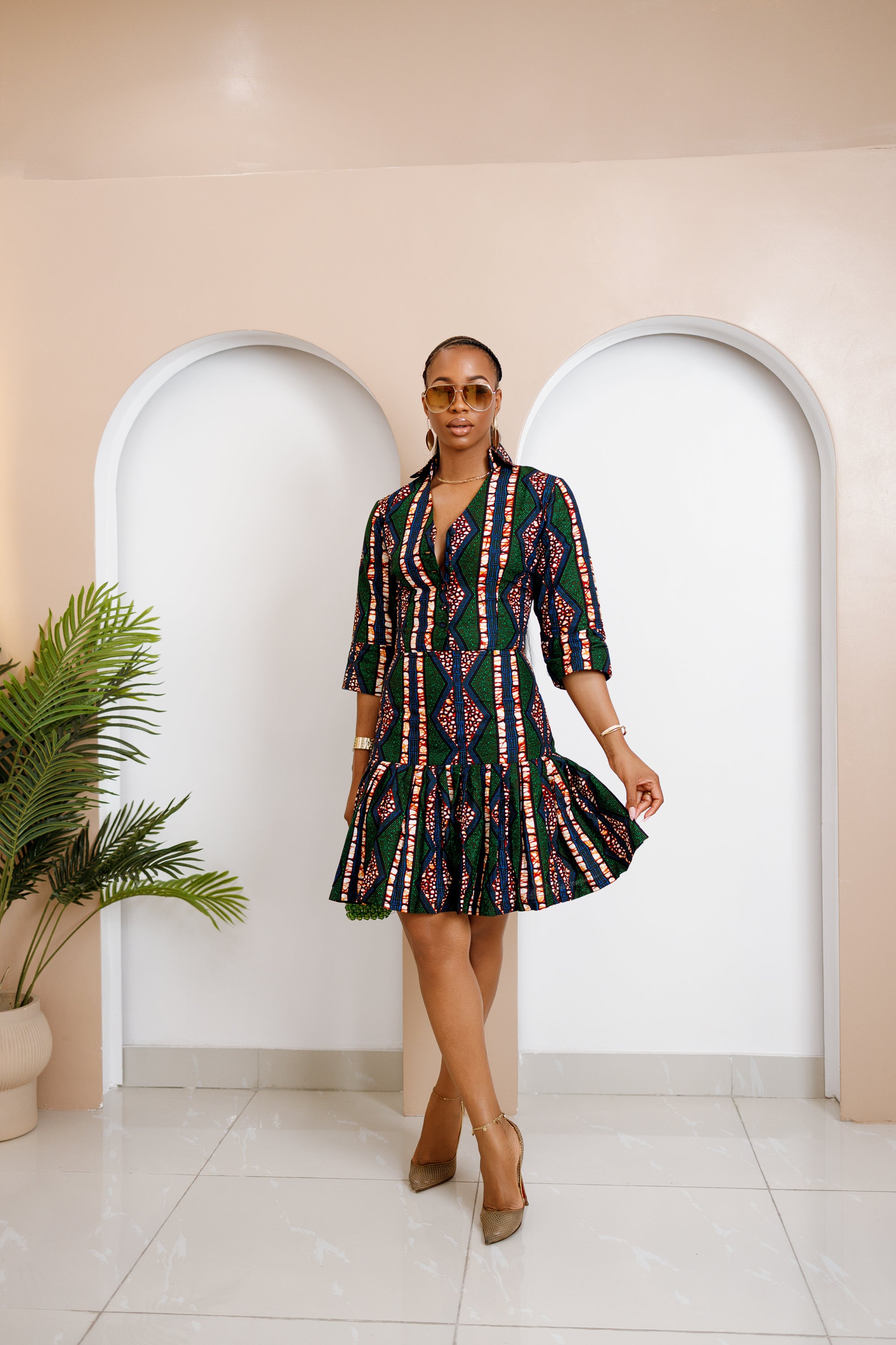 Model in Isiopko Ankara Midi Dress with structured bodice and pleated skirt in green, red, and blue African print, holding a green beaded handbag.