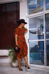 A woman posing in a yellow black and red two-piece set. She is pictured turned to the left with her hand on the wall behind her. She is styled with a black sun hat and silver jewelry.
