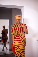 A woman posing in a yellow and red striped two-piece set. She is pictured facing the camera with her hand on her chest. She is styled with a brown leather hat and a red beaded purse.