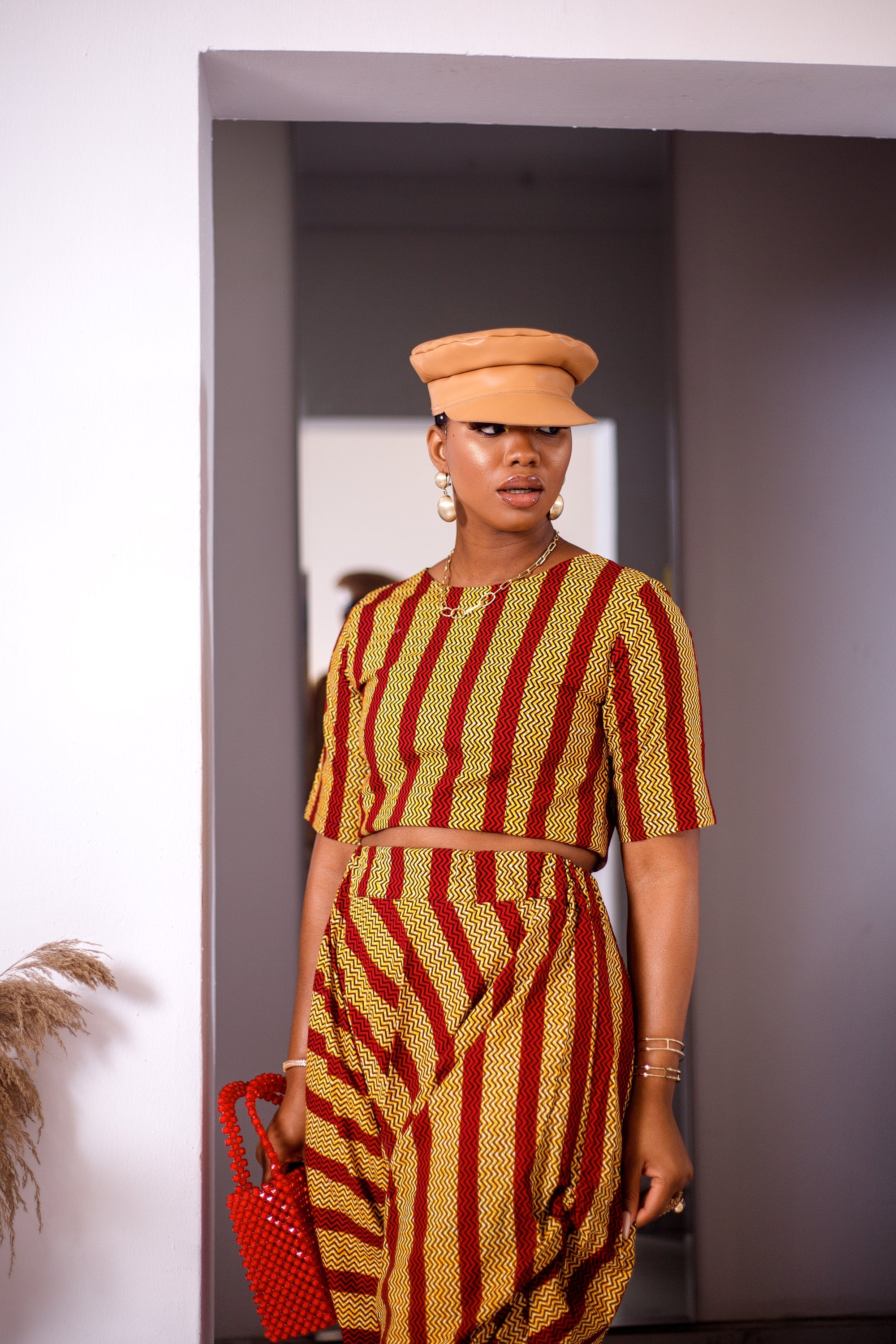 A woman posing in a yellow and red striped two-piece set. She is pictured facing the camera and looking to the side. She is styled with a brown leather hat and a red beaded purse.