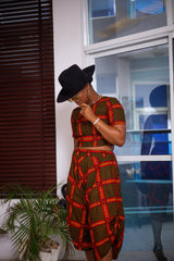A woman posing in a yellow black and red two-piece set. She is pictured facing the camera and looking down. She is styled with a black sun hat and silver jewelry.