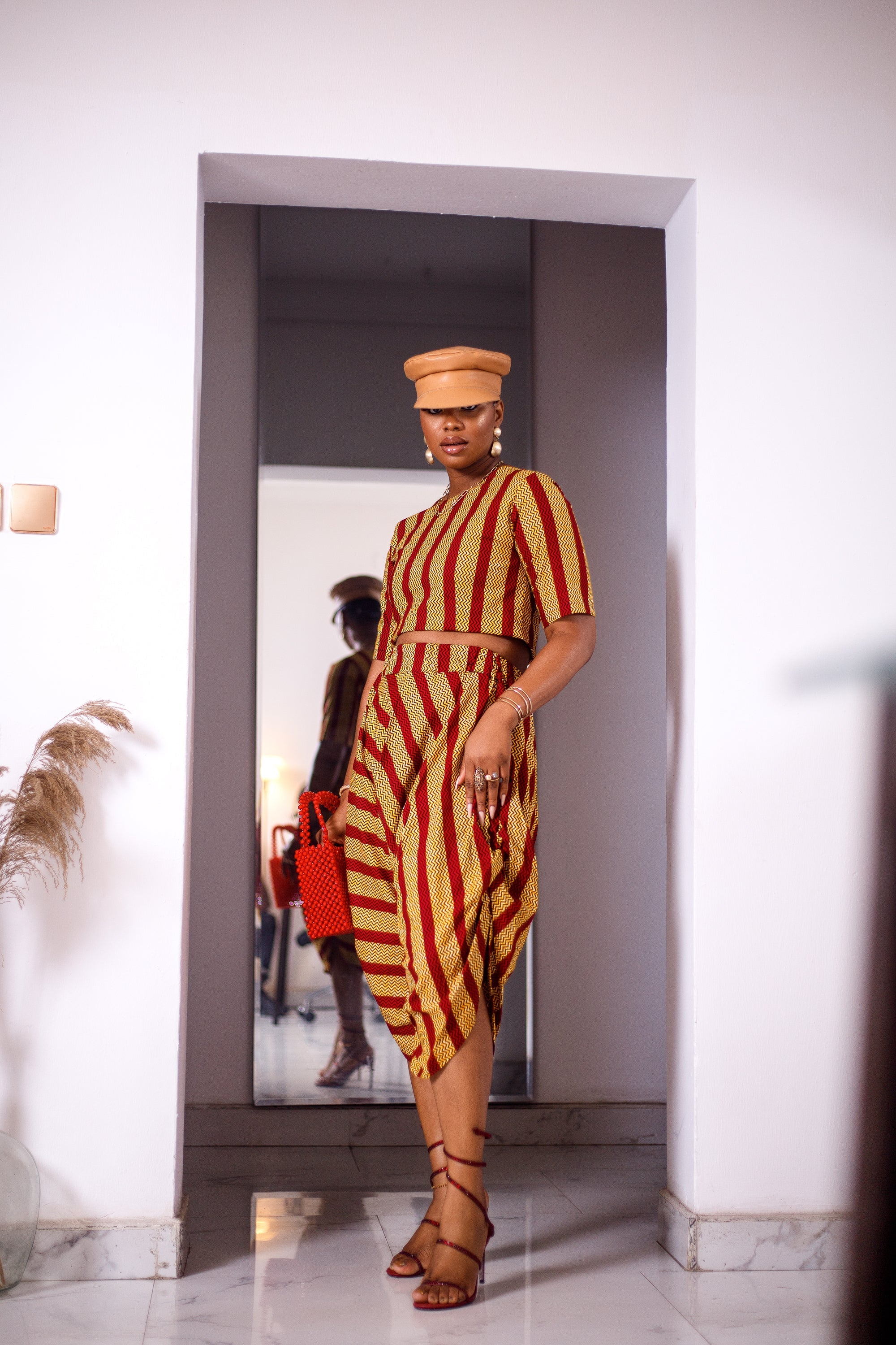 A woman posing in a yellow and red striped two-piece set. She is pictured facing the camera. She is styled with a brown leather hat and a red beaded purse.