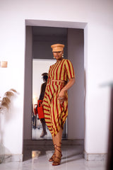 A woman posing in a yellow and red striped two-piece set. She is pictured facing the camera. She is styled with a brown leather hat and a red beaded purse.