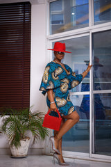 A woman posing in a blue and tan African print mini dress. She is pictured facing to the side with her right leg raised up. She is styled with a red beaded purse, a red sun hat, and silver jewelry.