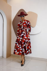 Woman wearing the Nwayioma Wrap Dress, a voluminous red African print wrap dress with pockets, styled with a wide-brim hat, sunglasses, and black heels.