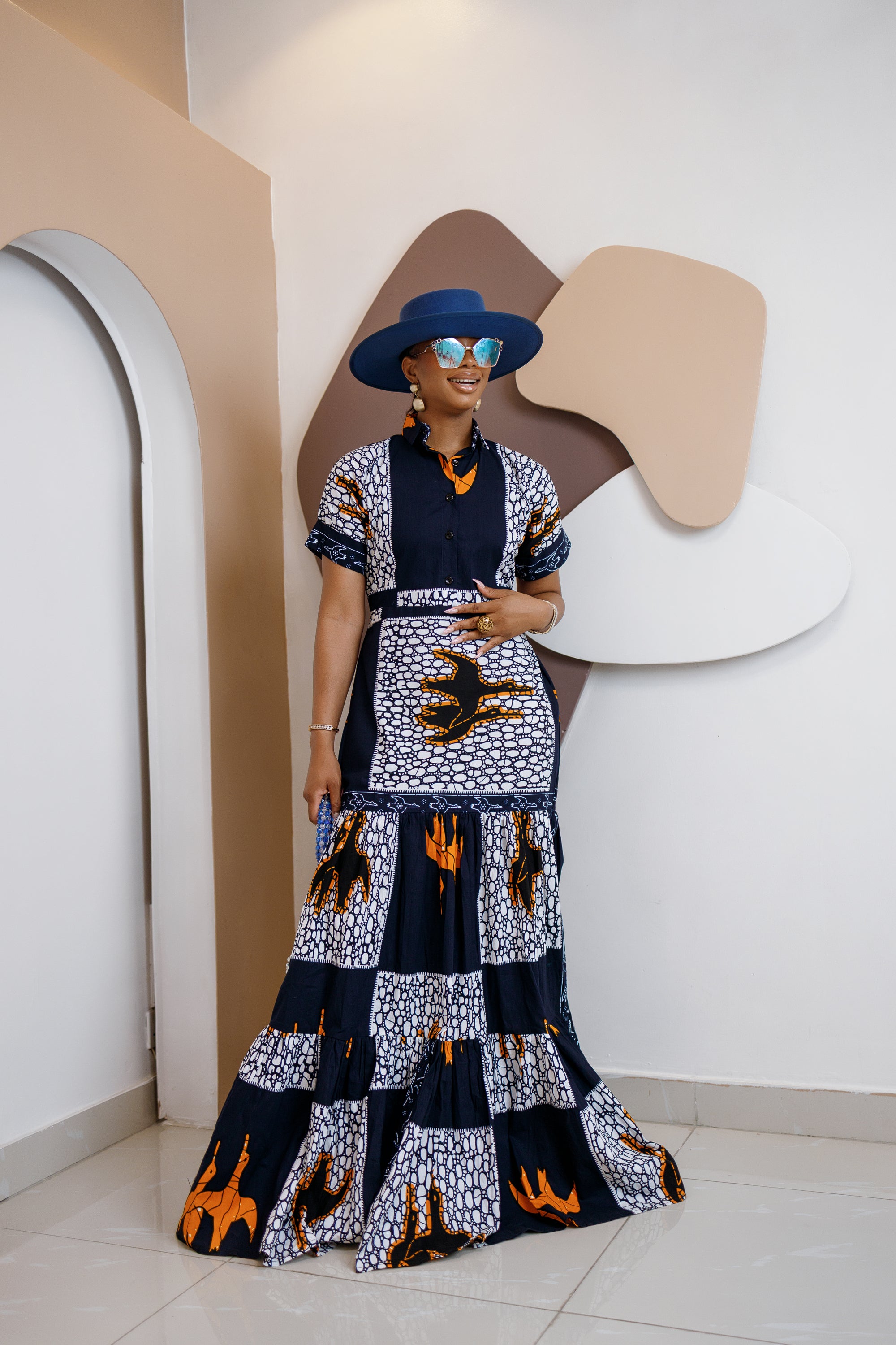 Model wearing Nzama Ankara maxi shirt dress with patchwork print in indigo, white, and orange, styled with a wide-brimmed hat and accessories.